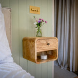 Wooden nightstand with a drawer and shelf against a light green wall with a curtain.