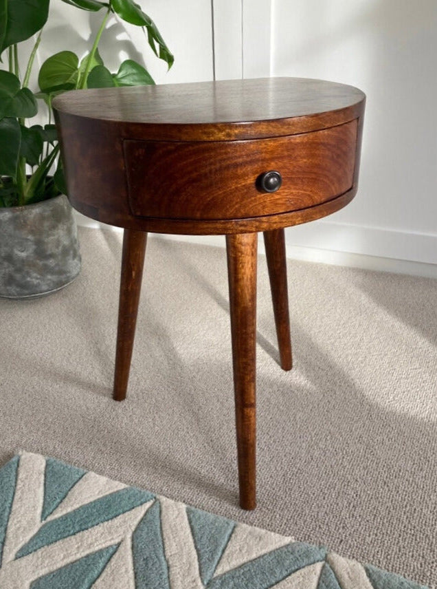 A semi circular shape bedside table made from mango wood in a dark brown finish, featuring a small drawer and single brass knob.