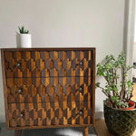 Wooden dresser with geometric pattern next to potted plants on a light-colored floor.