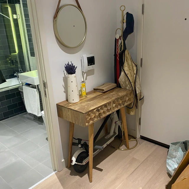 Natural mango wood console table with textured carved drawer fronts, in a room with a mirror, towel, and various items on a shelf.
