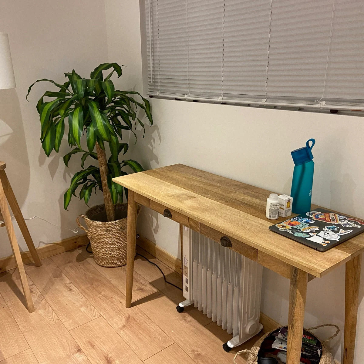 Wooden desk with items on a light wood floor next to a radiator and plant.
