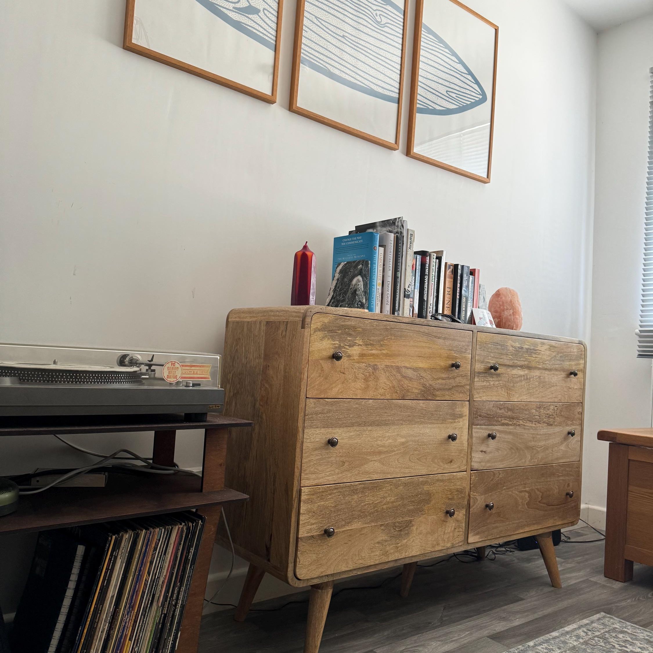 Curved chest of 6 drawers, made from mango wood, in a light brown finish and decorated with a row of books and a stylish salt lamp on top.