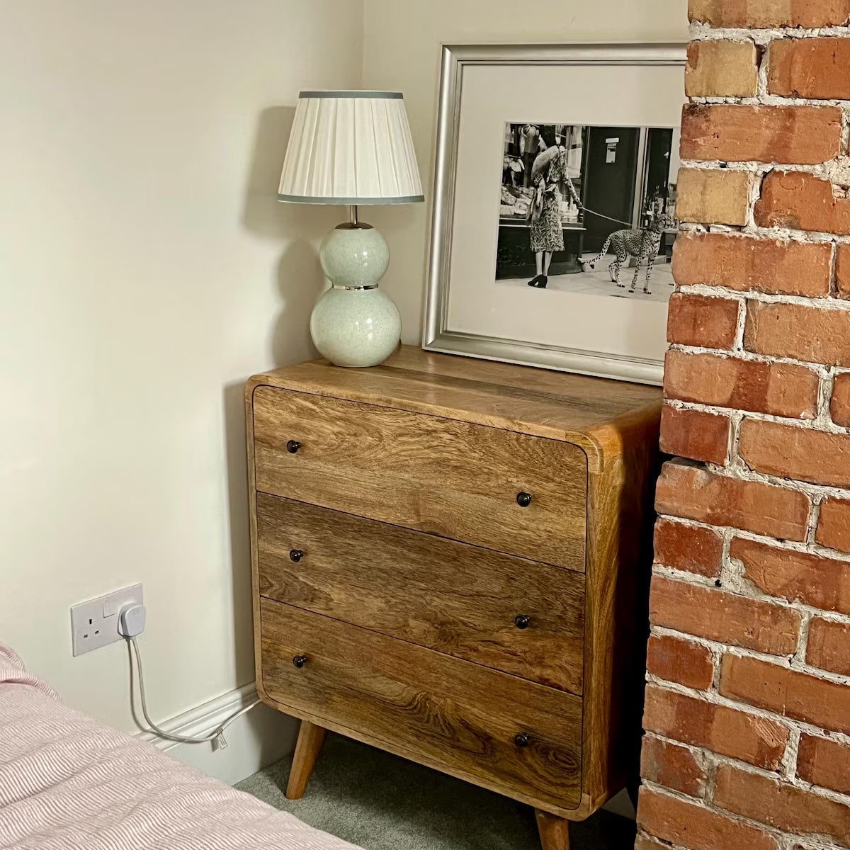 Wooden dresser with a lamp and framed picture against a brick wall.