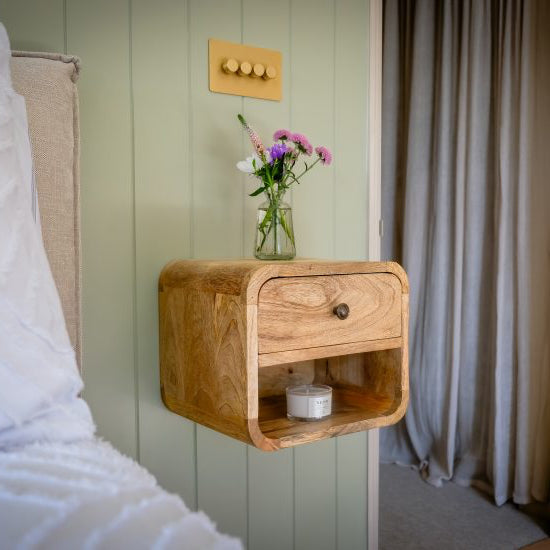 Wooden nightstand with a drawer and shelf against a light green wall with a curtain.