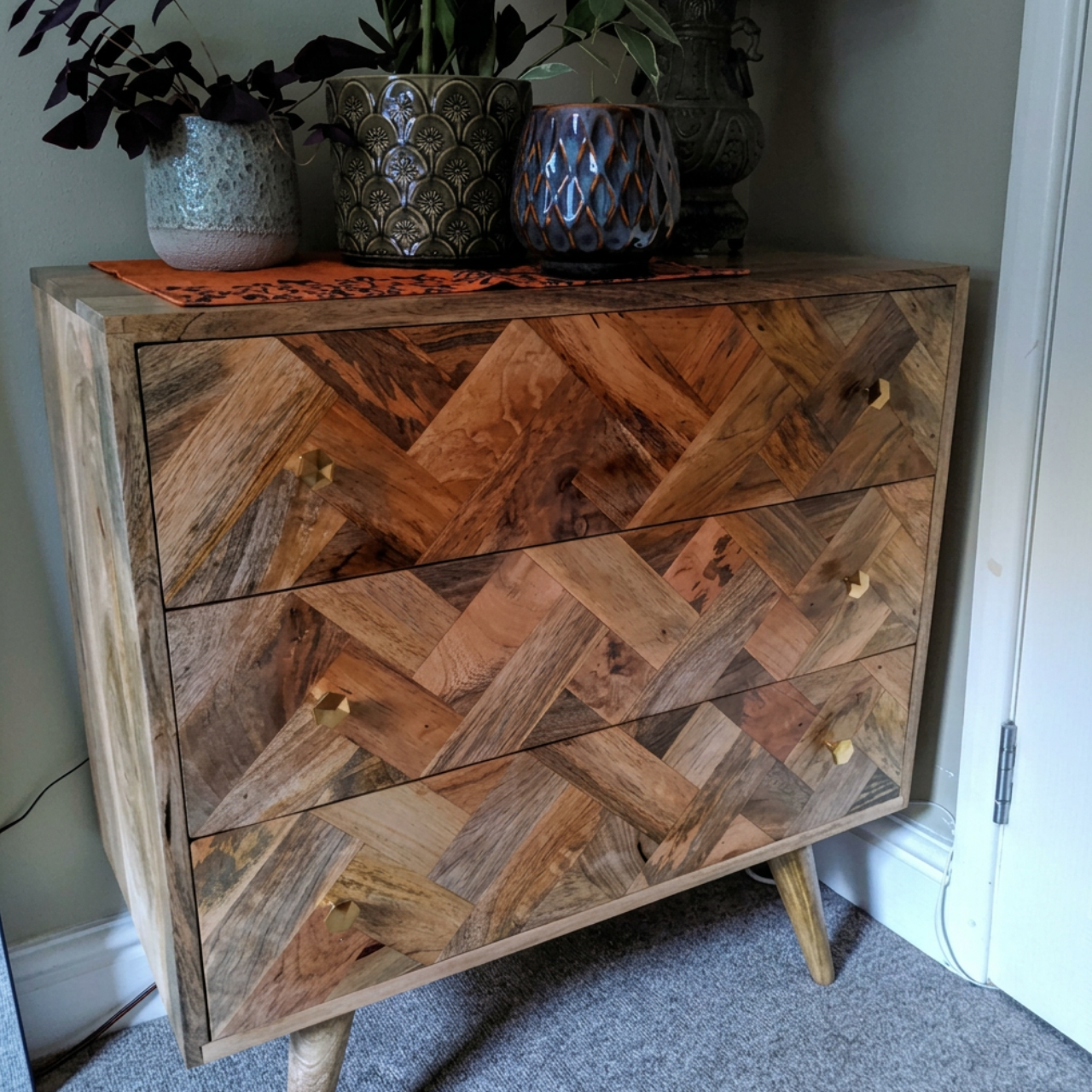 A mid-century modern wooden chest of drawers with a geometric herringbone pattern, styled with potted plants on top in a home setting.