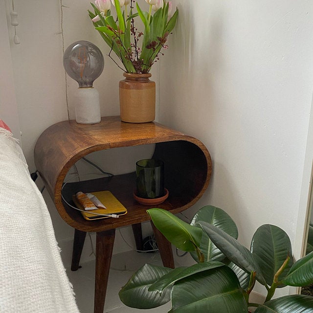A circular open bedside table, made from mango wood, in a dark, chestnut finish.