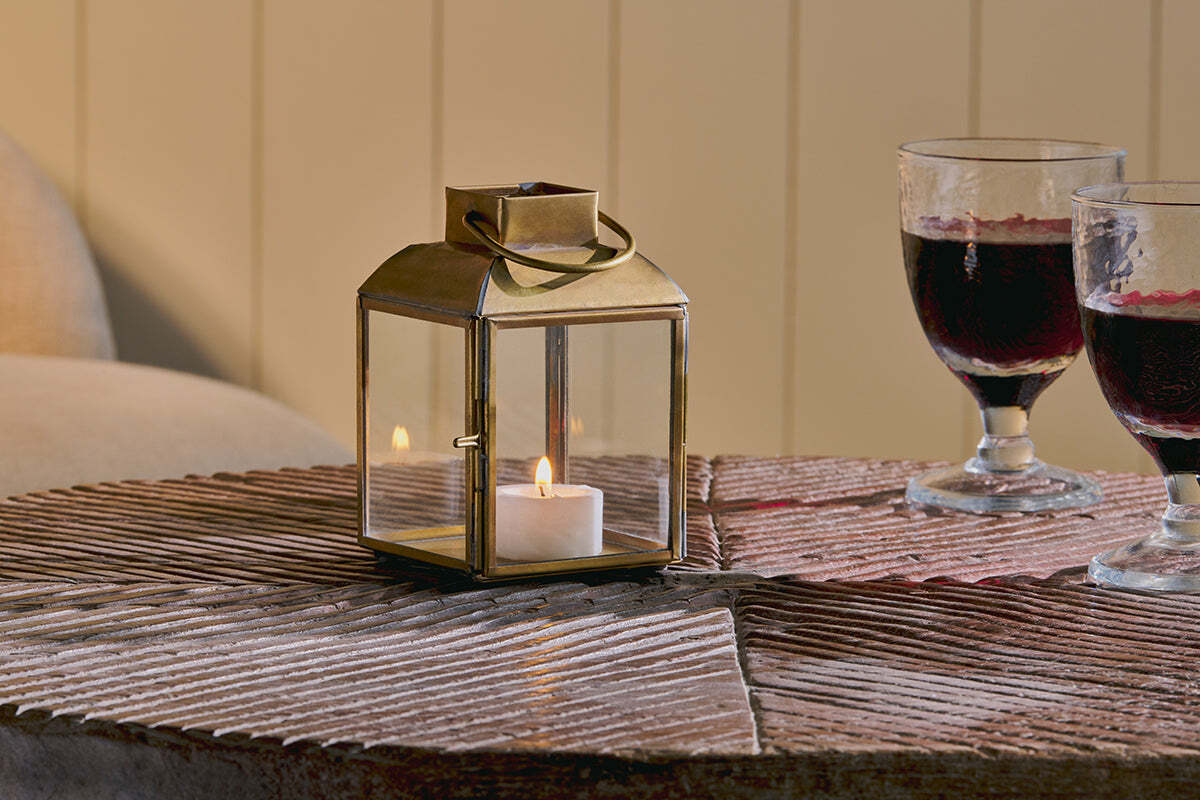 Candle lantern with a lit candle on a wooden surface next to two glasses of red wine.