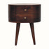 A round California walnut bedside table with two drawers and shiny brass knobs, perched on four tapered legs.
