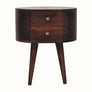A round California walnut bedside table with two drawers and shiny brass knobs, perched on four tapered legs.