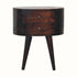 A round, dark brown finish mango wood bedside table with two drawers and four tapered legs, featuring brass knobs on the drawers.