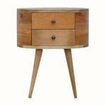 A round, natural finish mango wood bedside table with two drawers and four tapered legs, featuring brass knobs on the drawers.