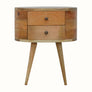 A round, natural finish mango wood bedside table with two drawers and four tapered legs, featuring brass knobs on the drawers.