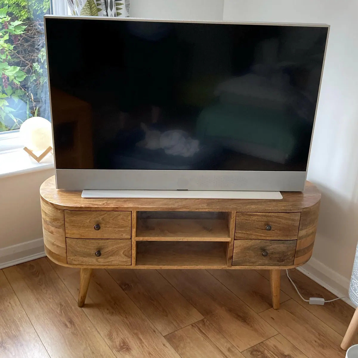 Large flat-screen TV on a wooden stand in a room with a window and plant.