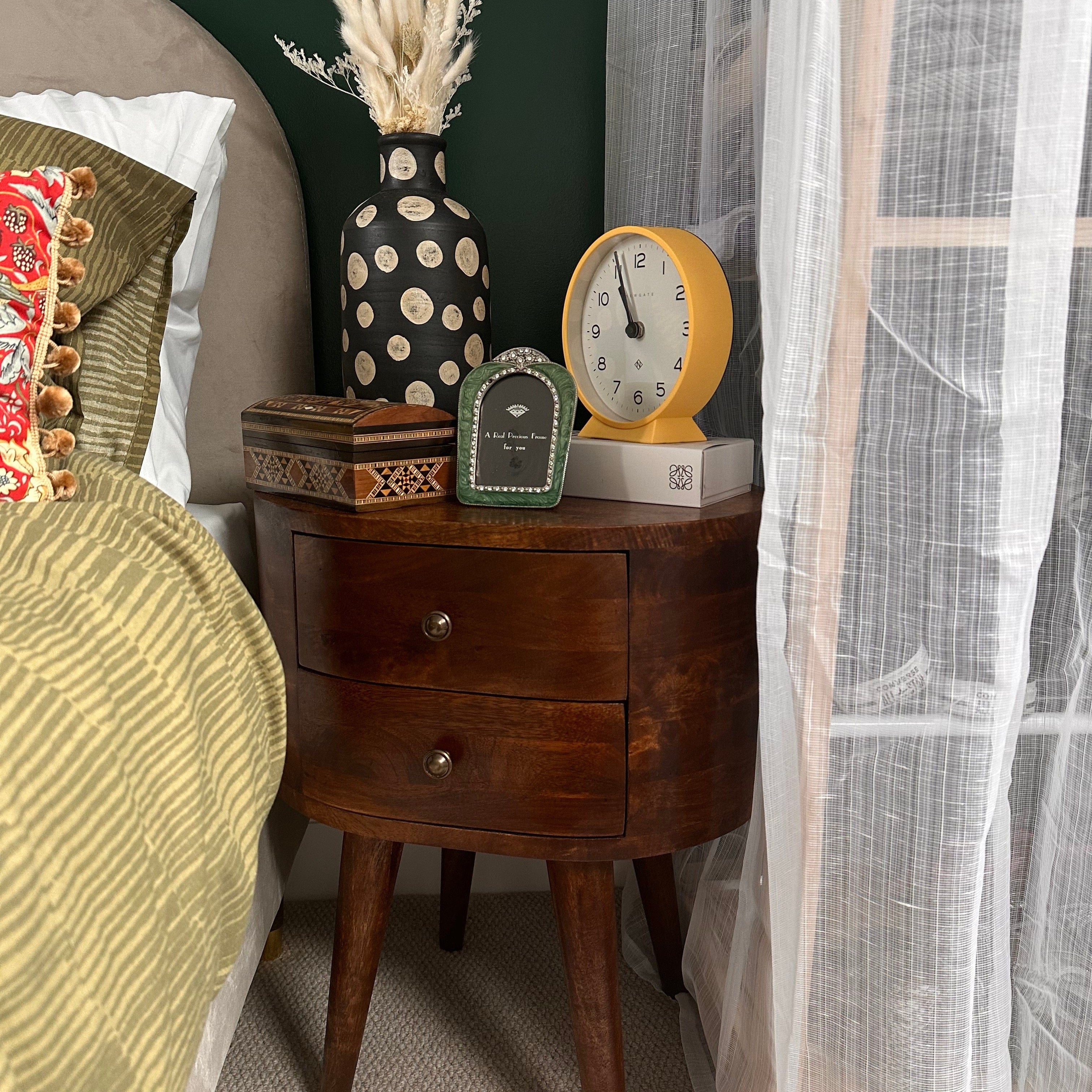 Bedroom featuring round chestnut-colored bedside table made of mango wood with two drawers and four tapered legs, featuring brass knobs.