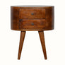 A round chestnut-colored bedside table made of mango wood with two drawers and four tapered legs, featuring brass knobs.