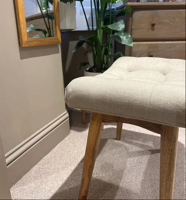 Beige cushioned stool with wooden legs in a room setting.